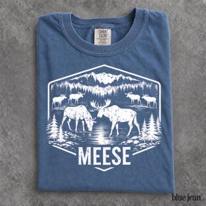 May include: A blue crew neck t-shirt featuring a white graphic print of a mountain landscape with a forest, a lake, and several moose. The text MEESE is printed in white capital letters at the bottom of the hexagonal graphic design.