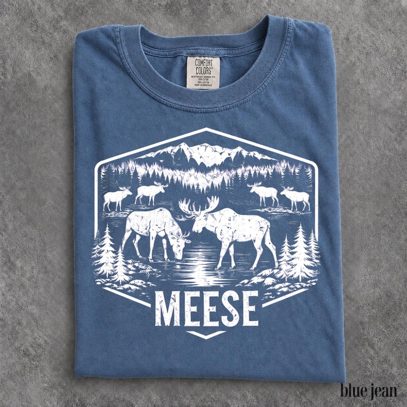 May include: A blue crew neck t-shirt featuring a white graphic print of a mountain landscape with a forest, a lake, and several moose. The text MEESE is printed in white capital letters at the bottom of the hexagonal graphic design.