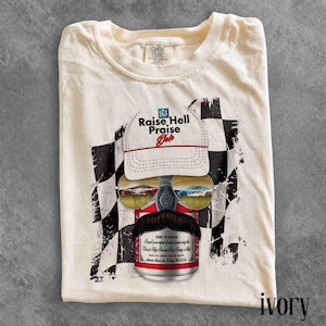 May include: An ivory t-shirt featuring a graphic of a white baseball cap with the text Raise Hell Praise Dale, sunglasses reflecting race cars, and a mustache above a red and white beverage can, all set against a black and white checkered flag.