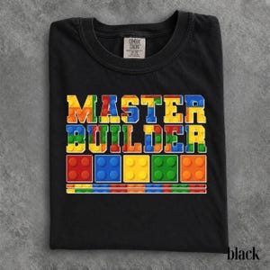 Può includere: Una t-shirt nera a girocollo con la scritta MASTER BUILDER in un carattere multicolore che ricorda i mattoncini da costruzione. Sotto il testo ci sono cinque icone di blocchi quadrati rossi, blu, gialli, verdi e arancioni con una linea.