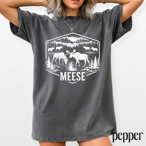 May include: A charcoal gray short-sleeve t-shirt featuring a white graphic print of a forest landscape with mountains, pine trees, and several moose. The text MEESE is printed in a bold white font below the central illustration of two moose.