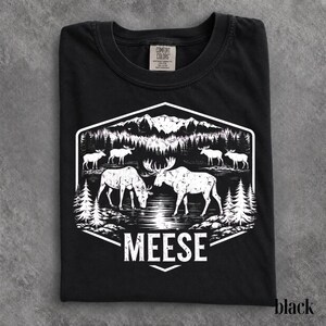 May include: A black t-shirt featuring a white graphic print of a mountain landscape with pine trees and several moose standing near a body of water. The word MEESE is printed in white capital letters at the bottom of the hexagonal design.