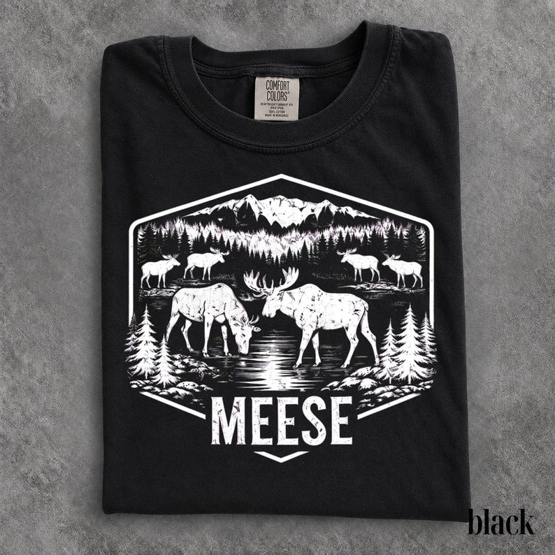 May include: A black t-shirt featuring a white graphic print of a mountain landscape with pine trees and several moose standing near a body of water. The word MEESE is printed in white capital letters at the bottom of the hexagonal design.