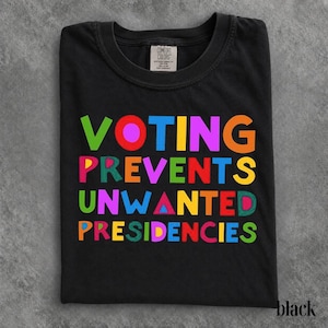 Puede incluir: Una camiseta negra de manga corta con cuello redondo y etiqueta de la marca Comfort Colors. El frente presenta el texto multicolor "VOTING PREVENTS UNWANTED PRESIDENCIES" en una fuente sans-serif audaz y divertida con letras en verde, rojo, azul, amarillo y rosa.