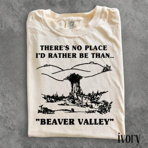May include: An ivory-colored short-sleeve t-shirt featuring a black graphic print. The text reads, "THERE'S NO PLACE I'D RATHER BE THAN..." above a line drawing of a landscape with hills and trees, followed by the text "BEAVER VALLEY" at the bottom.