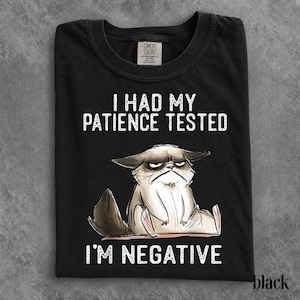 Op de afbeelding: Een zwart t-shirt met een afbeelding van een chagrijnige, zittende kat met een norse uitdrukking. De tekst boven de kat luidt I HAD MY PATIENCE TESTED en de tekst eronder luidt I'M NEGATIVE. Het t-shirt heeft een Comfort Colors merklabel.