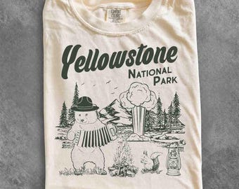 May include: An ivory t-shirt featuring a dark green graphic print with the text Yellowstone National Park. The design depicts a bear wearing a hat and playing an accordion, a campfire, a geyser, a squirrel, and a lantern amidst pine trees.
