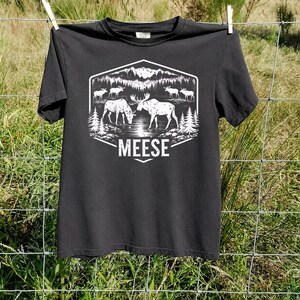 May include: A black short-sleeved t-shirt featuring a white graphic print of a forest landscape with mountains, pine trees, and several moose. The text MEESE is printed in a bold, white sans-serif font below the central illustration.