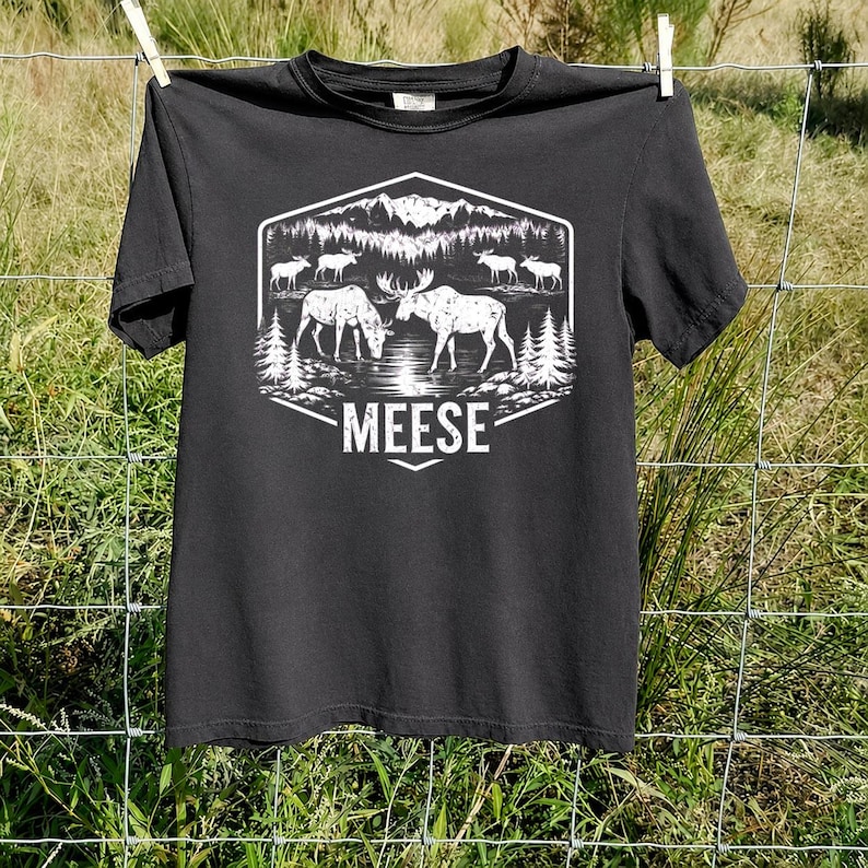 May include: A black short-sleeved t-shirt featuring a white graphic print of a forest landscape with mountains, pine trees, and several moose. The text MEESE is printed in a bold, white sans-serif font below the central illustration.