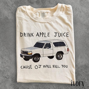 May include: A cream-colored short-sleeve t-shirt featuring a graphic of a white SUV. The text above the vehicle reads DRINK APPLE JUICE and the text below reads CAUSE OJ WILL KILL YOU. The shirt has a Comfort Colors brand label at the neckline.