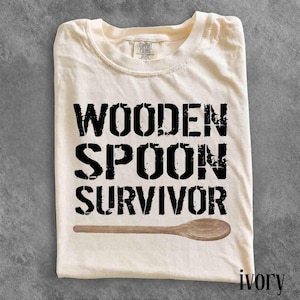 May include: An ivory-coloured short-sleeve t-shirt featuring the black text "WOODEN SPOON SURVIVOR" in a distressed stencil font, with a graphic illustration of a brown wooden spoon positioned horizontally underneath the text.