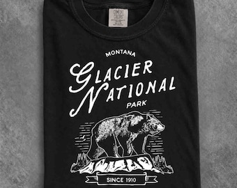 May include: A black t-shirt featuring a white graphic print of a bear standing on a rocky landscape. The text reads MONTANA GLACIER NATIONAL PARK and SINCE 1910. The shirt has a crew neckline and a Comfort Colors brand label on the inner collar.