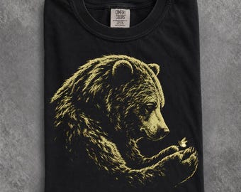 May include: A black crew neck t-shirt featuring a graphic print of a bear in profile, rendered in a light yellow hue. The bear is looking down at a small bee resting on its paw. The inner neck label displays the text Comfort Colors 100% Cotton.