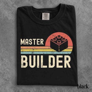 Puede incluir: Una camiseta negra de cuello redondo con un estampado gráfico que incluye el texto MASTER BUILDER en una fuente desgastada. El diseño presenta un icono de bloque de construcción negro sobre un motivo de atardecer semicircular con rayas horizontales en naranja, amarillo y turquesa.