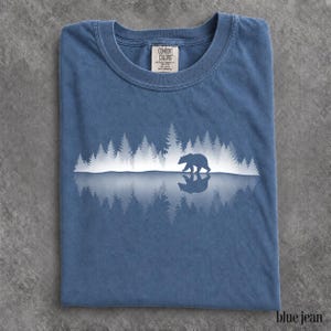 May include: A blue short-sleeve t-shirt featuring a white graphic of a bear walking through a forest with a mirrored reflection below. The inner neck label reads Comfort Colors, 100% cotton, made in Honduras. The fabric has a washed, textured appearance.