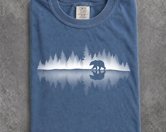 May include: A blue short-sleeve t-shirt featuring a white graphic of a bear walking through a forest with a mirrored reflection below. The inner neck label reads Comfort Colors, 100% cotton, made in Honduras. The fabric has a washed, textured appearance.