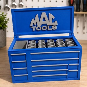 May include: A blue tool chest organizer with a hinged lid featuring the text "MAC TOOLS" in raised silver lettering. The interior contains a grey rectangular compartment and multiple grey cylindrical slots for tool storage, with silver drawer pulls.
