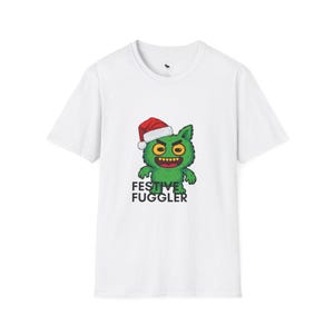 May include: A white short-sleeve t-shirt featuring a graphic of a green furry creature wearing a red and white Santa hat. The creature has large yellow eyes and a wide grin with visible teeth. Below the graphic is the text FESTIVE FUGGLER.
