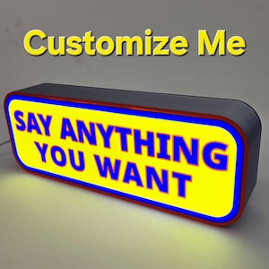May include: A rectangular LED light box with rounded corners, featuring a bright yellow face with a blue border and red trim. The text "SAY ANYTHING YOU WANT" is printed in bold blue letters on the front, with "Customize Me" above it.
