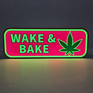 May include: A rectangular LED sign with rounded corners featuring a vibrant pink background and a bright green border. The text WAKE & BAKE is displayed in bold green capital letters alongside a green cannabis leaf graphic on the right side.