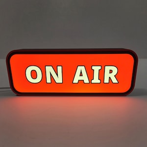 May include: A rectangular illuminated sign with rounded corners featuring a bright orange background and the text ON AIR in bold white capital letters. The sign has a dark frame and is designed to be displayed as a broadcast indicator light.