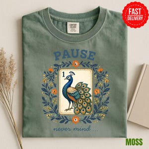 May include: A sage green t-shirt featuring a graphic of a peacock on a rectangular card labeled with the number 1. The peacock is surrounded by a circular wreath of blue leaves and orange flowers. The text PAUSE is above and never mind is below.
