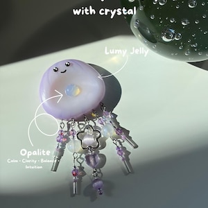 Op de afbeelding: Een lichtpaarse Lumy Jelly bedel met een lachend gezicht en een ingebed opaliet kristal. De bedel heeft bungelende zilverkleurige kettinkjes versierd met paarse en doorzichtige kralen, een bloemvormige bedel en een hartvormige kraal.
