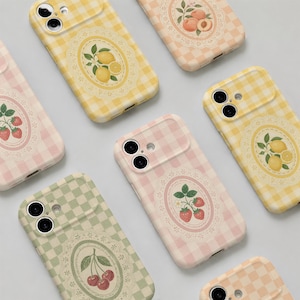 May include: A collection of smartphone cases featuring a chequered pattern in various pastel colours. Each case displays a central oval frame with a lace-like border containing a detailed illustration of fruit, including strawberries, lemons, and peaches.