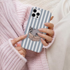 Könnte beinhalten: Eine Smartphone-Hülle mit einem Muster aus vertikalen hellgrauen und weißen Streifen. In der Mitte befindet sich eine verspielte Illustration eines kleinen grauen Waschbären, der in einem braunen Pappkarton sitzt, im einfachen Linienstil.