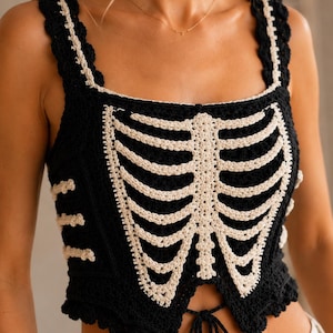 May include: A black crochet tank top featuring a cream-colored rib cage skeleton design on the front. The garment has thick straps, a square neckline, and a scalloped hem with a small tie closure at the center of the waist for a fitted look.
