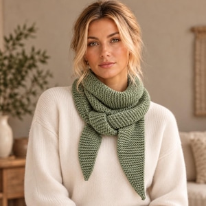 Può includere: Una sciarpa lavorata a maglia color verde salvia con un motivo a legaccio testurizzato, avvolta attorno al collo in stile triangolare. La sciarpa presenta estremità appuntite e una struttura a maglia grossa, indossata sopra un maglione color crema.