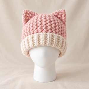 May include: A crochet beanie hat in a soft pink colour featuring two pointed cat-like ears on top. The hat has a thick, ribbed cream-coloured brim at the base and a textured, chunky knit pattern throughout the main body of the headwear piece.
