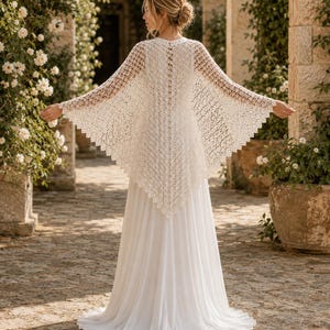 Può includere: Un abito bianco lungo fino ai piedi con un coprispalle in uncinetto color crema a maniche lunghe e orlo a punta. Il coprispalle presenta un intricato motivo a maglia aperta che si estende dalle spalle ai polsi e lungo la schiena.