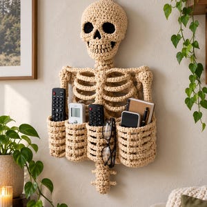 May include: A beige crochet wall organizer shaped like a human skeleton torso with a skull head. The rib cage section features four integrated pockets holding various items including remote controls, a smartphone, a notebook, a pen, and eyeglasses.