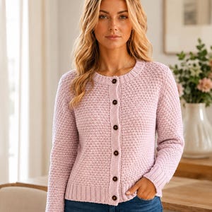 May include: A light pink knit cardigan featuring a textured waffle-stitch pattern, a round neckline, and a front button closure with dark brown buttons. The garment has long sleeves with ribbed cuffs and a ribbed hemline, providing a structured fit.