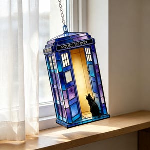 May include: A hanging stained glass lamp shaped like a blue police box with the text POLICE PUBLIC CALL BOX. The lamp features a warm interior light and a small black cat silhouette sitting inside the open doorway, suspended by a metal chain.