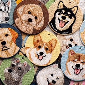 Op de afbeelding: Een verzameling ronde geborduurde patches met diverse hondenrassen, waaronder een husky, poedel, corgi en shiba inu. Elke patch toont een gedetailleerd geborduurd portret van een hondenhoofd in levendige kleuren op een effen achtergrond.