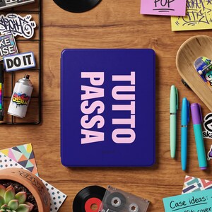 May include: A rectangular dark blue electronic reader case featuring the light pink text TUTTO PASSA printed vertically in a bold sans-serif font. The case has a smooth, solid-coloured surface and is positioned centrally in a flat-lay composition.