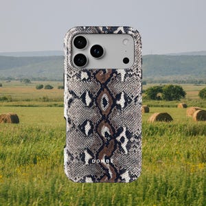 May include: A rectangular smartphone case featuring a textured snakeskin pattern in shades of brown, black and white. The case has a cutout for a triple-lens camera system and the text DOMBE printed in white on the lower centre section.