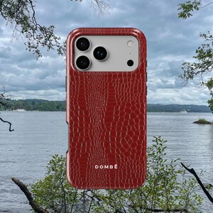 Puede incluir: Una funda de smartphone roja con un patrón texturizado de piel de cocodrilo y un área blanca para la cámara. La palabra DOMBĒ está impresa en blanco cerca de la parte inferior central de la funda. Incluye tres lentes circulares y un flash.