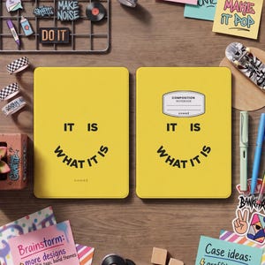 May include: Two bright yellow notebooks featuring the black text IT IS WHAT IT IS arranged in a smiley face pattern. The right notebook includes a white composition notebook label. Surrounding items include colourful sticky notes, graffiti pins, and tape.