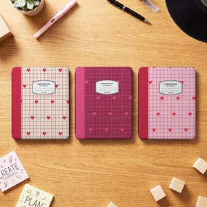 Puede incluir: Tres cuadernos de composición con un patrón de cuadrícula y pequeños corazones rojos. El cuaderno de la izquierda es crema, el central es magenta oscuro y el de la derecha es rosa claro. Cada uno tiene un lomo de color sólido y una etiqueta blanca.