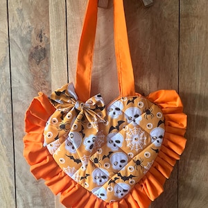 May include: A heart-shaped tote bag with orange fabric handles and a ruffled orange border. The bag features a quilted pattern with a print of white skulls, black spiders, brown bats, and orange eyes on a golden yellow background, plus a bow.