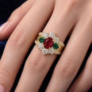 May include: A gold ring featuring a central oval-cut red ruby surrounded by two pear-cut green emeralds and six round-cut white diamonds. The wide band is accented with small pavé-set diamonds along the edges, creating a cluster design.