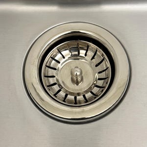 Puede incluir: Un colador circular de acero inoxidable para fregadero de cocina con acabado metálico pulido. El colador cuenta con un pomo central para levantarlo y una serie de ranuras verticales espaciadas uniformemente en un patrón radial.