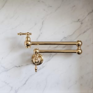 May include: A polished gold-tone wall-mounted pot filler faucet featuring a double-jointed swing arm, a circular base plate, and two lever handles. The faucet has a metallic finish with a reflective surface and a classic, multi-part construction.