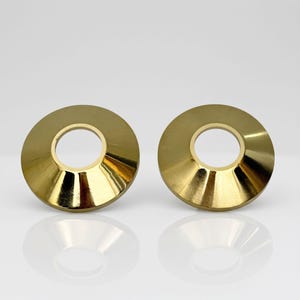 Könnte beinhalten: Zwei kreisförmige, goldfarbene Abdeckrosetten aus Metall mit einer glatten, reflektierenden Oberfläche. Jedes Teil verfügt über einen breiten, abgeschrägten Außenrand, der sich zu einer mittigen, runden Öffnung verjüngt und einheitlich wirkt.