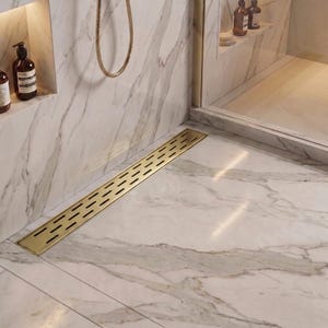 Könnte beinhalten: Ein rechteckiger, gebürsteter goldener linearer Duschablauf mit einem Muster aus horizontalen Schlitzen, der bündig in einen weißen Duschboden aus Marmor mit grauer Maserung eingelassen ist, nahe einer Glasduschwand und Wandseifenspendern.