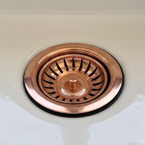 Puede incluir: Un colador de desagüe para fregadero de cobre pulido con un borde circular y un pomo central. El colador presenta una serie de ranuras curvas y alargadas dispuestas en un patrón radial, sobre una superficie de lavabo blanca lisa.