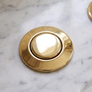 以下が含まれることがあります： 光沢のあるゴールドトーンの金属製シンク排水栓。滑らかで反射する表面と中央の盛り上がったボタンデザインが特徴です。この金具は、繊細なグレーの筋が入った白い大理石の表面に取り付けられており、清潔感のあるメタリックな仕上がりを見せています。